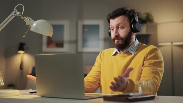 Shrug, i dont know. Perplexed bearded man with headphones in home office looking at computer webcam and emotionally squeezes his shoulders