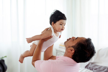 Obraz na płótnie Canvas Asian single dad plays with newborn son in bedroom. He lifted his son up and pretended to be flying in the air with joy and fun. Concept Happy Family