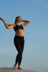 Young woman in sportswear with long hair standing on the rock by the sea and enjoying sunrise on a summer morning. Blue sky on the background.