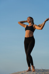 Obraz premium Young woman in sportswear with long hair standing on the rock by the sea and enjoying sunrise on a summer morning. Blue sky on the background.