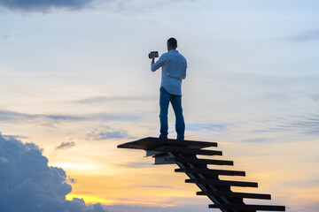 Obraz premium A man in jeans and a white shirt is standing on the stairs against the colorful sky with a camera in his hand