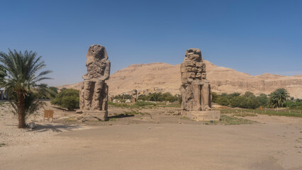 Giant statues of the colossi of Memnon against the blue sky and sand dune. Huge sculptures of...