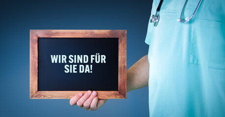 Wir sind für Sie da!. Arzt zeigt Schild/Tafel mit Holz Rahmen. Hintergrund blau