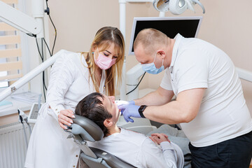 Obraz premium The dentist doctor looks at the patient's teeth and holds dental instruments near the mouth. The assistant helps the doctor. They wear white uniforms with masks and gloves. Dentist. Dental office