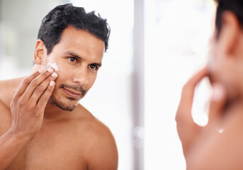 He takes great care of his skin. A young man applying cream to his face while looking in the mirror.