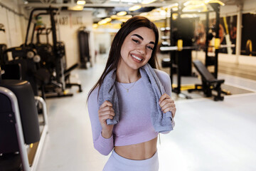 Fototapeta premium Fit young woman with a towel at health club. Female relaxing after workout. Portrait of charming smiling young fitness girl with a towel and posing while looking at the camera in the gym.