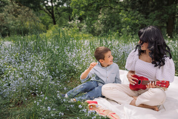 Fototapeta premium Mother singing playing on ukulele while sitting with her son on picnic blanket in the park.