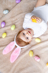 Obraz na płótnie Canvas cute little baby with bunny ears and painted eggs lies on a beige background and smiles, easter concept