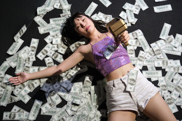Young asian woman surrounded by dollar bills and point to the camera with a toy gun.