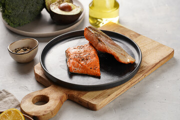 Two grilled fish steaks - red salmon on black round plate on wooden board, olive oil, avocado on grey background