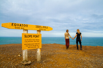 Slope point signpost and back view couple of travelers. The southernmost point of South Island New Zealand. South Pacific Ocean