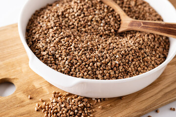 dry buckwheat groats in a white bowl on a wooden board with a wooden spoon, on a white background, top view, close-up