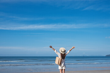 Rear view image of a woman with hat and bag opening arms while walking on the beach with blue sky background