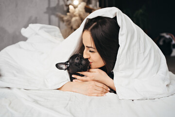 Beautiful brunette woman with a French Bulldog puppy in a bed under a blanket.