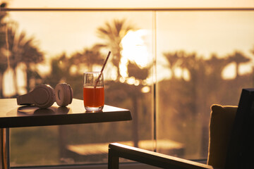 The glass of juice and headphones on the table on balcony opposite sunset palm sea beach. Rear view