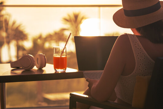 Silhouette of woman freelancer in straw hat drinking juice during working on laptop sitting on the armchair near table on balcony opposite sunset palm sea beach. Remote work on the vacation. Rear view