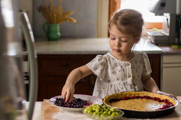 Portrait of little blonde cheerful girl decorating a pie fruits and berries on a wooden table in the kitchen on the background of light window