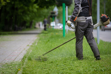A worker in protective clothing with a trimer in his hands, mows the grass along the road. The powerful mower mows flowers and other plants. Territory care.