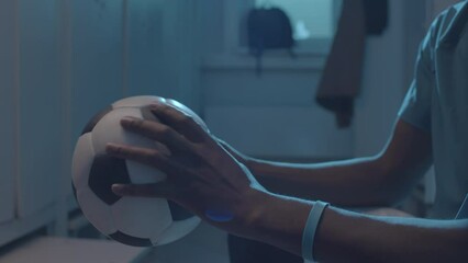 Midsection of cropped male athlete sitting in locker room, tossing soccer ball