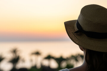 Silhouette of woman in straw hat standing on balcony and looking at sunrise palm sea beach. Rear...