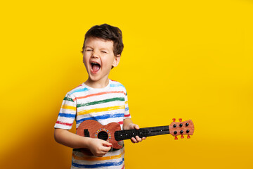 funny boy playing guitar. A child with a guitar on a yellow background in the studio. Emotions. The...