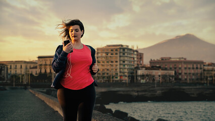 Beautiful chubby young woman in sportswear with wired earphones running seaside and looking on the...