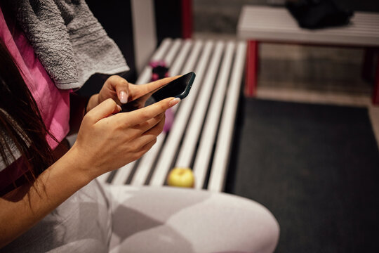 Young athletic woman using mobile phone after sports training at gym dressing room. Tired female athlete using smart phone on a break after sports training at gym locker room.