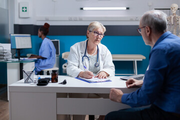 Medic signing clipboard document and doing medical consultation with patient. Woman doctor writing checkup papers to give prescription medicine to senior man at healthcare appointment.