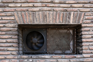 light red brick wall with a small window one part of which has a built-in large old fan with a rusty metal..