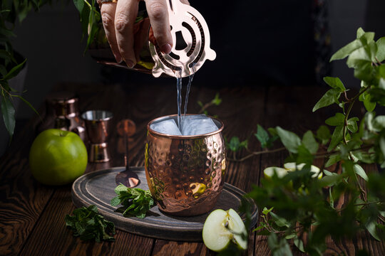 Woman hand pouring Green Apple Irish Mule cocktail cocktail from shaker in copper mug surrounded by ingredients and bar tools