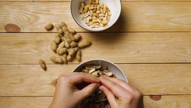 Close-up of women's hands peeling peanuts over bowls. A handful of peanuts lie on a wooden table. Top view. Peeling an earthy peanut at home