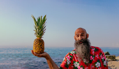 photo of a shaved head man with long beard dressed with hawaiian shirt holding a pineapple on the beach with a surprised expression in his face - Powered by Adobe