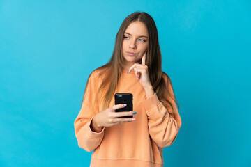 Young Lithuanian woman isolated on blue background using mobile phone and thinking