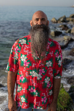 waist up portrait image of a hawaiian shirt dressed bearded man with bald head standing in a beach enjoying the summer with unfocused stones and waves in the background