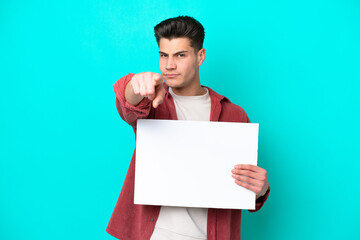 Young handsome caucasian man isolated on blue bakcground holding an empty placard and pointing to the front