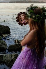 Mavka water nymph naiad near the water in nature forest on the river bank of the lake on a quarry on a cliff in a purple lilac transparent dress with a wreath flying mirror Ivan Kupala