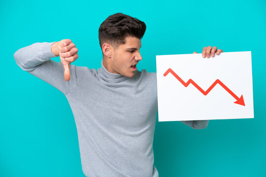 Young handsome caucasian man isolated on blue bakcground holding a sign with a decreasing statistics arrow symbol and doing bad signal