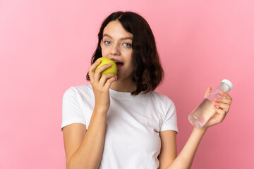 Teenager Ukrainian girl isolated on pink background with a bottle of water and eating an apple