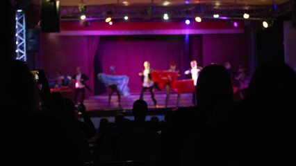 Blurred musical show on stage, dancers in colorful costumes and tailcoats performing cancan in slow motion, viewed from behind heads of audience. Concept of entertainment