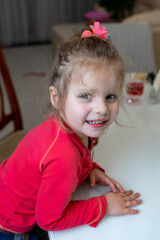 Charming little girl with blonde hair and colorful hairpins looks at the camera, smiles sweetly and feels good at table in the kitchen