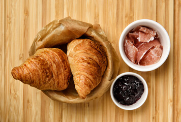 Fresh baked croissants on in basket with baking paper lining, parma ham and blueberry jam in small white bowl on wooden board