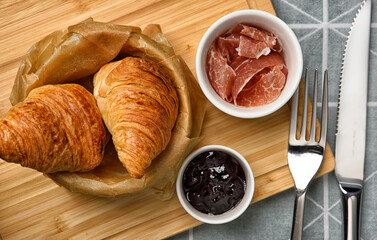 Fresh baked croissants on in basket with baking paper lining, parma ham and blueberry jam in small...