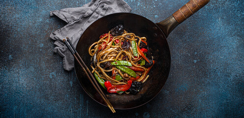 Asian dish stir fry udon noodles with vegetables and mushrooms in black rustic wok pan with wooden chopsticks on rustic dark blue concrete background from above, Chinese fast food