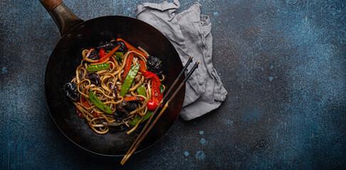 Asian dish stir fry udon noodles with vegetables and mushrooms in black rustic wok pan with wooden...