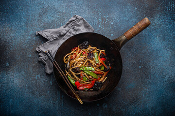 Asian dish stir fry udon noodles with vegetables and mushrooms in black rustic wok pan with wooden chopsticks on rustic dark blue concrete background from above, Chinese fast food