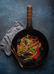 Asian dish stir fry udon noodles with vegetables and mushrooms in black rustic wok pan with wooden...
