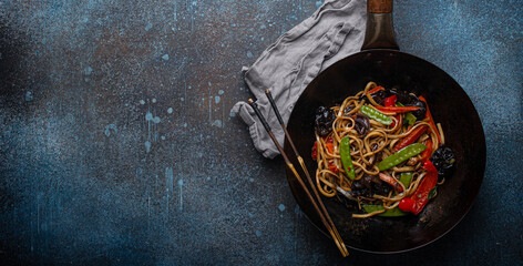 Asian dish stir fry udon noodles with vegetables and mushrooms in black rustic wok pan with wooden...