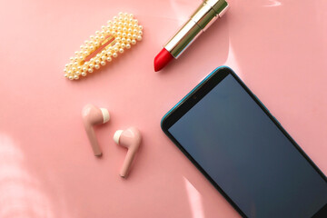 Mobile phone, pink wireless earphones, red lipstick and pearl hair clip on pastel pink background....