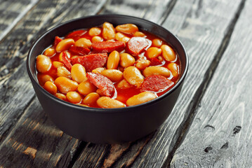 Beans baked with sausages. Beans with fried sausages in tomato sauce. Food in a black bowl on a brown wooden table.