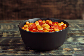 Beans baked with sausages. Beans with fried sausages in tomato sauce. Food in a black bowl on a brown wooden table.